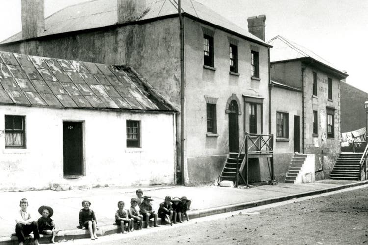 This black and white historical photo shows a small group of children sitting on the kerb in front of some houses. The images appears to have been taken in the late 18000s or early 1900s.