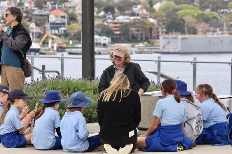 A small group of students sitting down infront of a smiling teacher. In the background is water and a distant suburb.