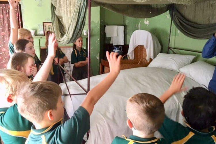 A group of students with hands raised to answer a question standing around an historical bed. The walls are green and the bed is covered by a canopy.