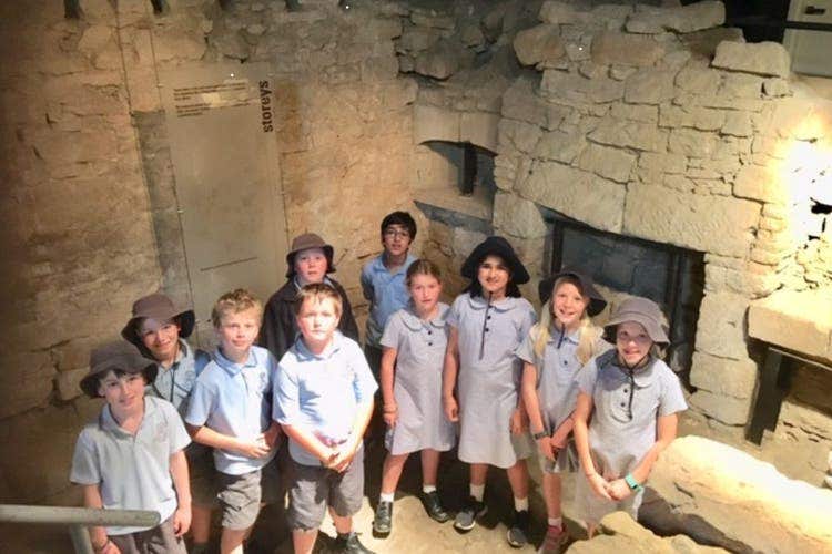 A group of students in the ruins of an old convict house. The students are stood, in a semi circle, and behind them is the remenants of an old sand stone wall, inset is a fireplace.