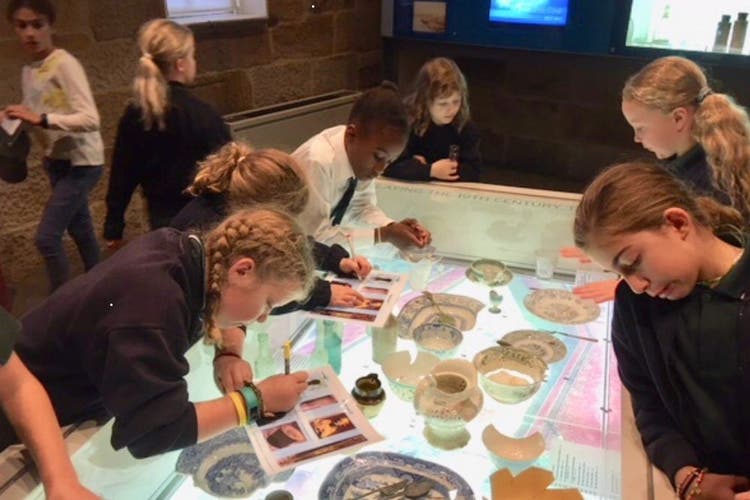 A group of students gathered around a collection of artefacts. Students are recording their findings on their worksheets.
