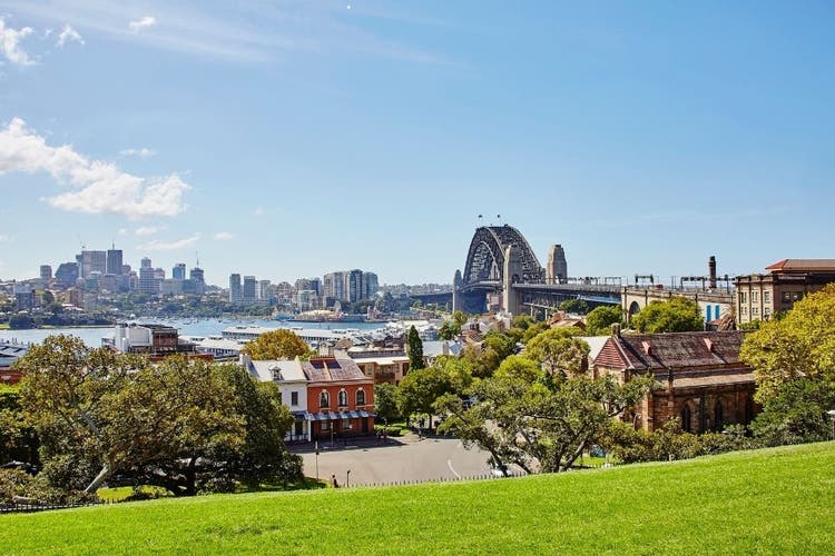 Sydney Harbour Bridge sits in the background, along with the North Sydney skyline. The photograph is taken from Observatory in. The sky is blue and there is a lawn with lush green grass.