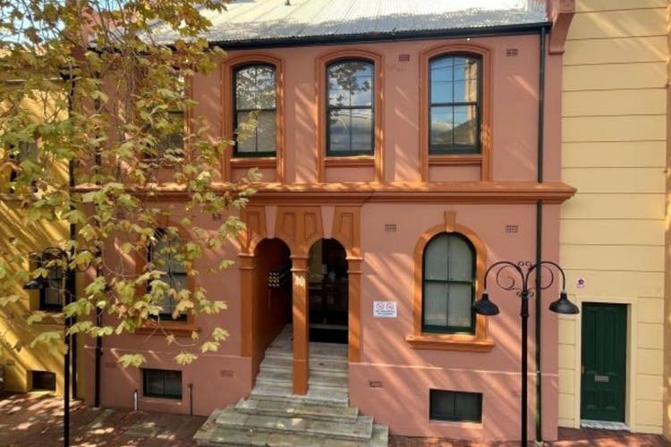A pinkish coloured building that sits to the left a yellow coloured one. A tree partially obscures the view of the building. The doors, in the centre, are both arches.